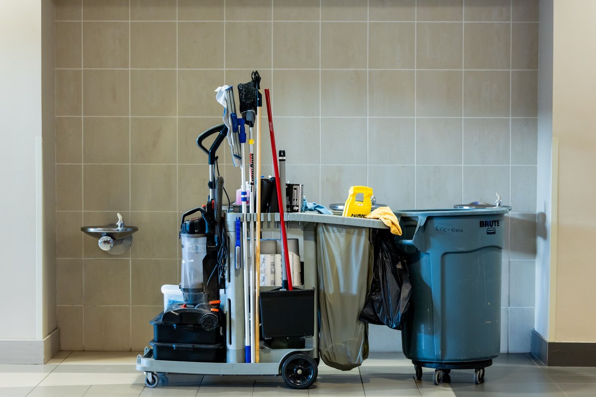 Janitorial cart loaded with a vacuum, mops, supplies and a gray trash bin in a tiled commercial hallway