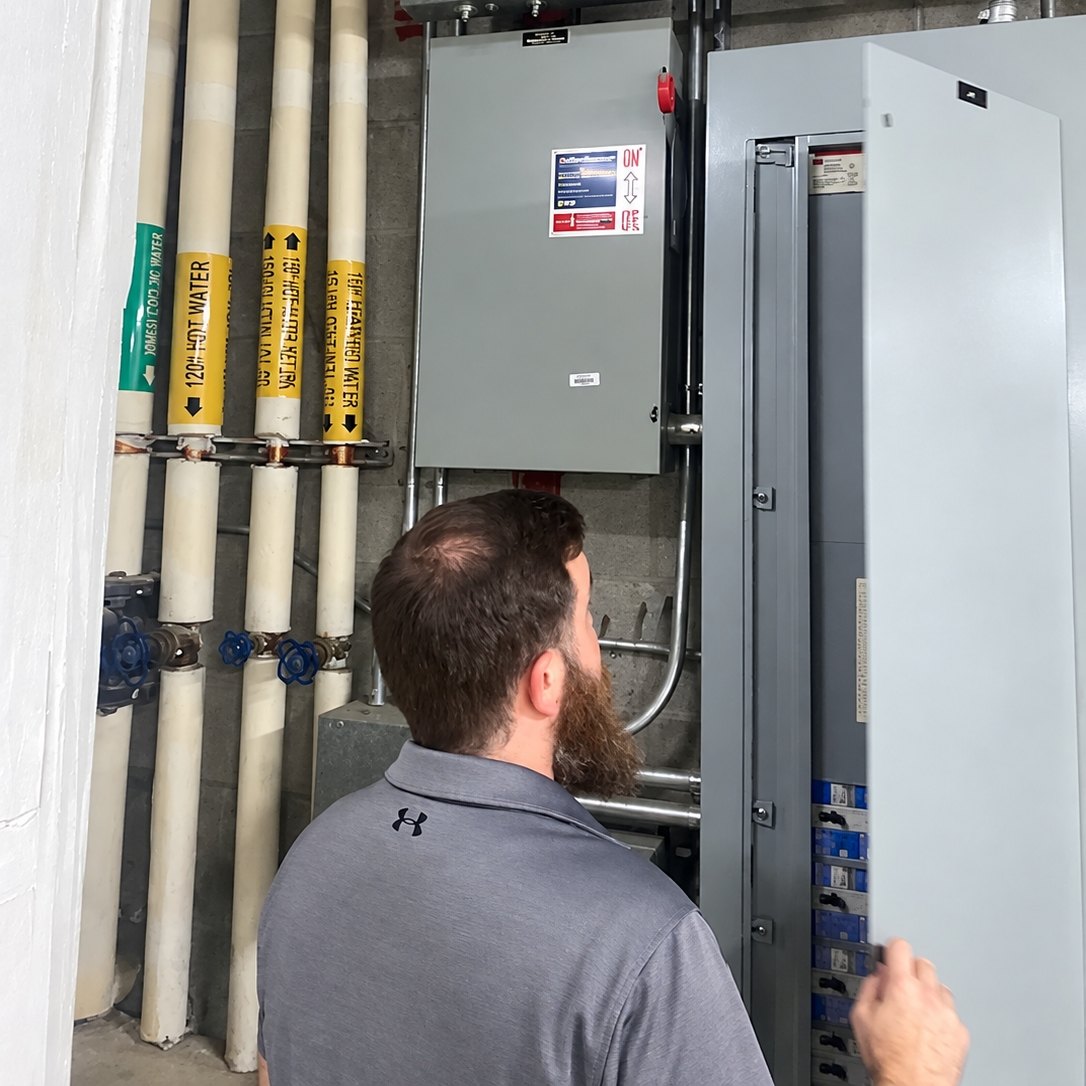 Technician in a gray polo opening a commercial electrical panel beside labeled hot water pipes