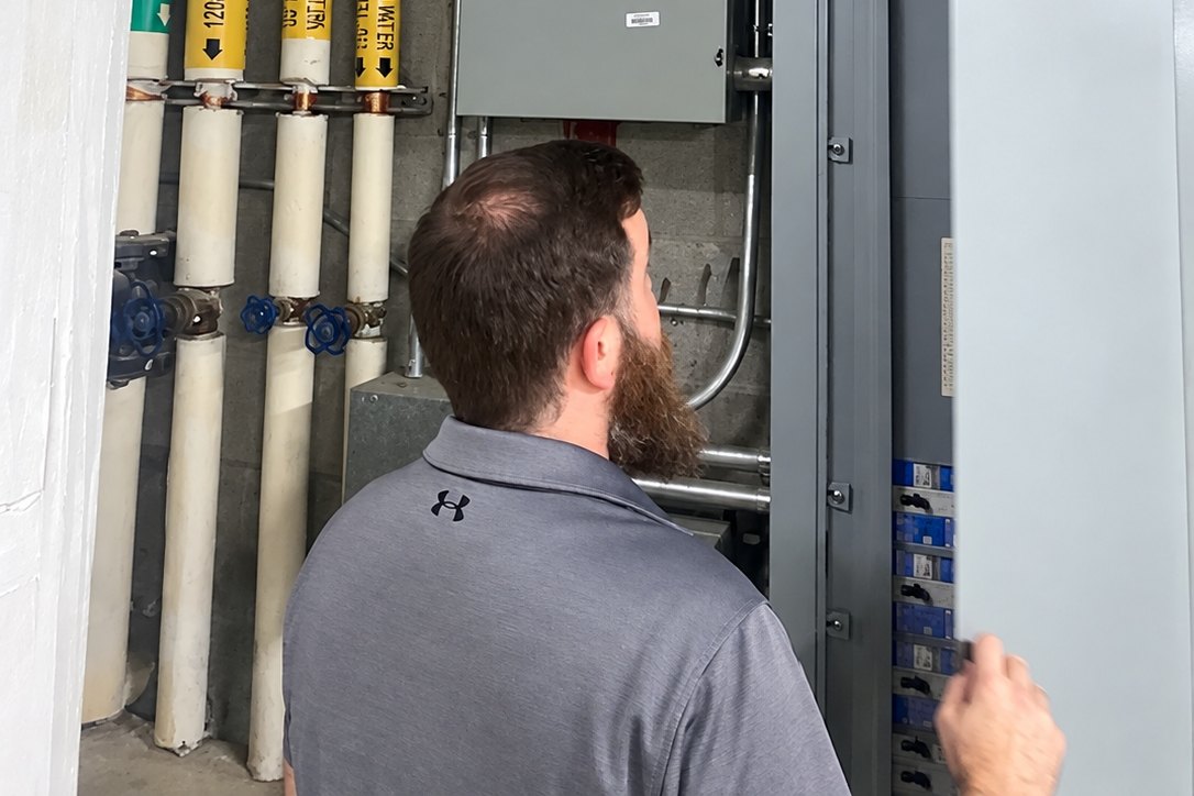 Technician opening a tall commercial electrical panel beside labeled hot water pipes