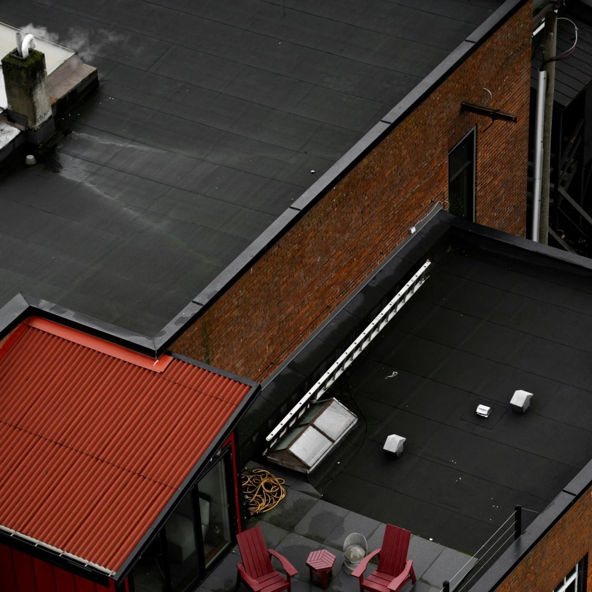 Aerial view of flat black roofs, a red corrugated roof section, a brick wall, a ladder and roof vents