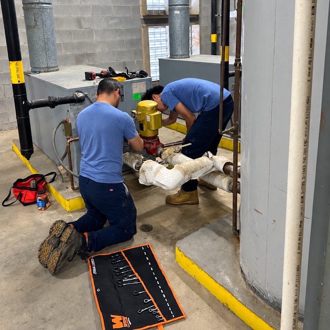 Two technicians kneeling at a yellow circulator pump on insulated piping beside a gray boiler