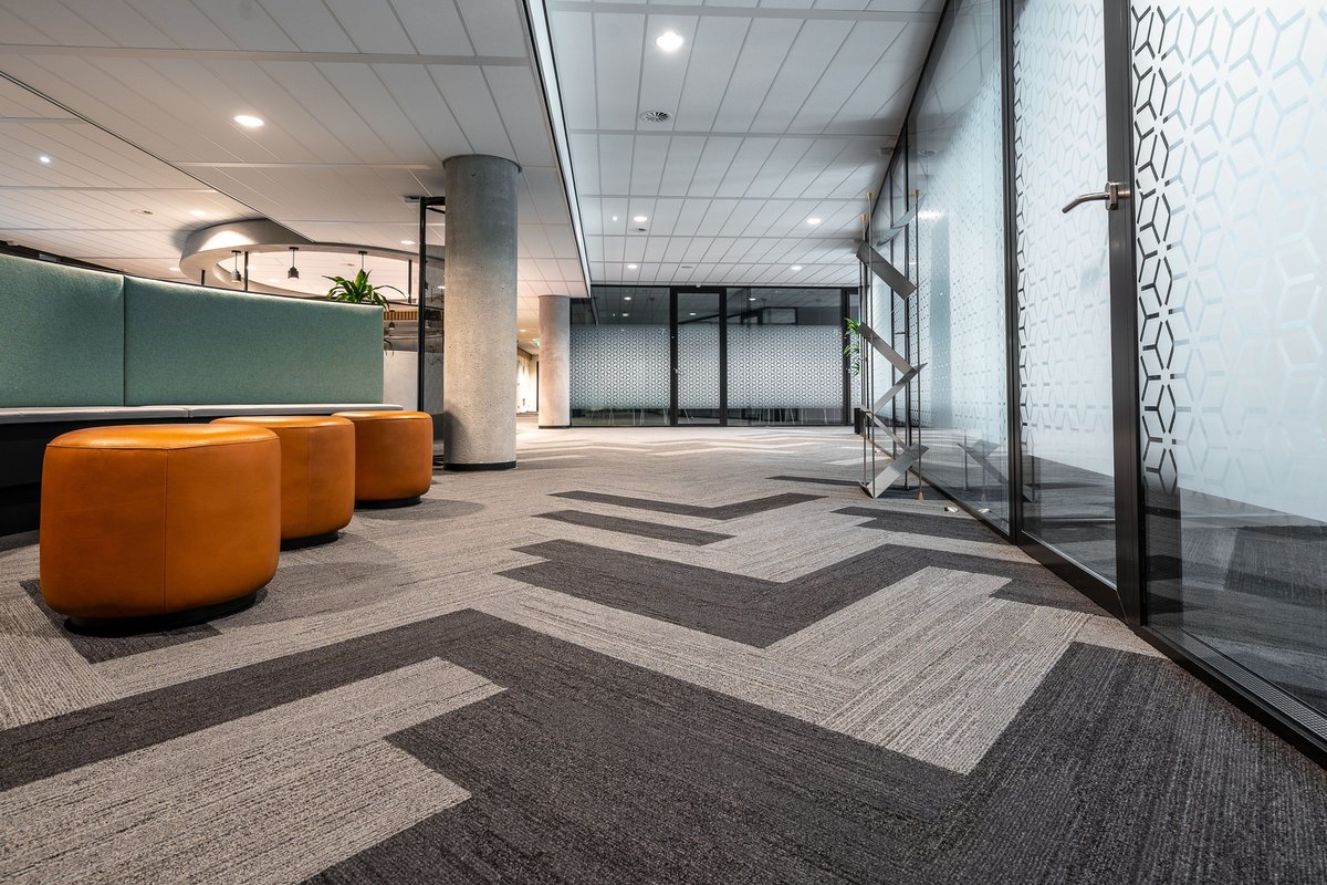 Office corridor with a gray herringbone carpet floor, orange leather ottomans and frosted glass walls