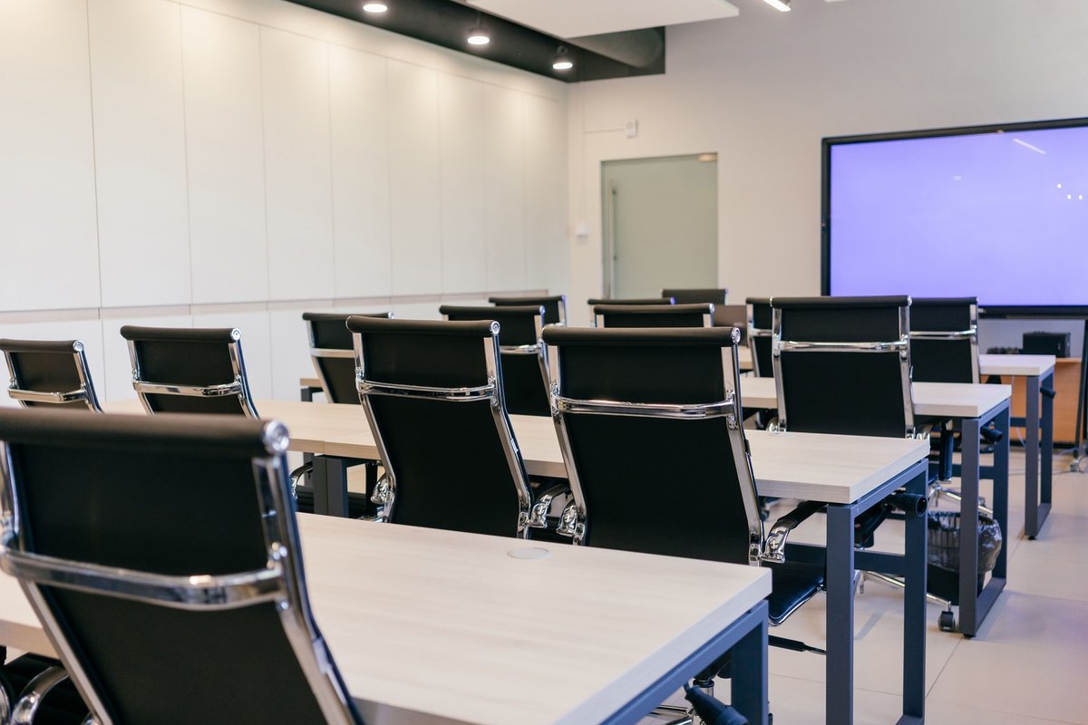 Bright meeting room with rows of wood-top desks, black leather chairs and a large wall screen